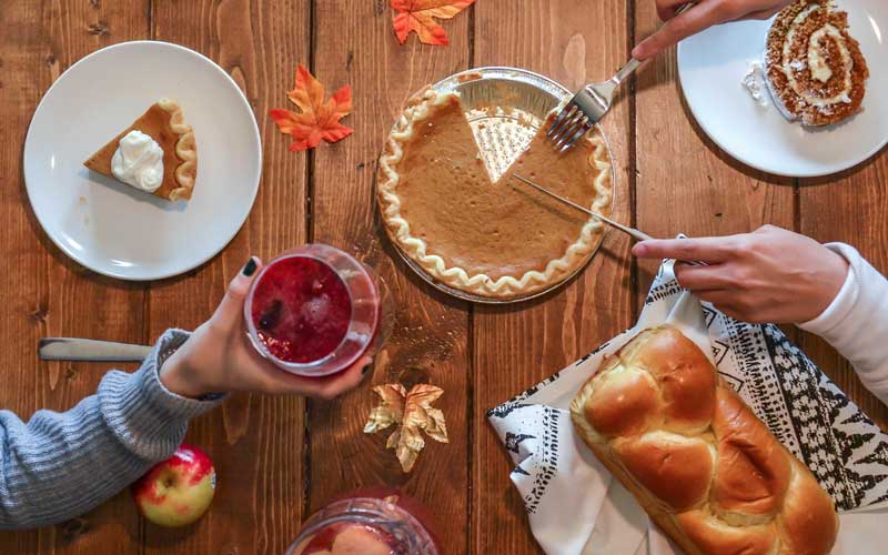 Two people serve and enjoy pumpkin pie, braided bread, and drinks at a wooden table decorated with autumn leaves.